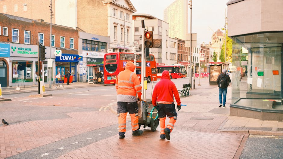 Two street cleaners wearing orange and red high-visibility uniforms are walking on a pedestrian crossing in Maida Vale, London, pushing a wheeled cleaning machine. They are surrounded by a busy urban environment with shops, including a Yorkshire Building Society branch, and a red double-decker bus in the background. The scene is well-lit with natural daylight, showcasing an outdoor city setting with paving stones and a glass storefront on the right. Cleaner Maida Vale ensures thorough surface cleaning and sanitation in residential and commercial spaces, highlighting professional cleaning services aligned with the focus on hygiene and maintenance.