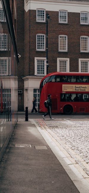 Street scene in Maida Vale featuring a red double-decker bus parked beside a brick building with multiple white-framed sash windows. Pedestrians are walking along the pavement, which includes a smooth concrete sidewalk and cobblestone road, illuminated by natural daylight. The building's exterior showcases a mix of red and brown brickwork, and the ground floor appears to be part of a commercial or retail space with glass windows and minimal clutter. The setting emphasizes urban cleanliness and maintenance, consistent with domestic cleaning services offered by Cleaner Maida Vale, including surface cleaning and sanitisation of accessible areas.