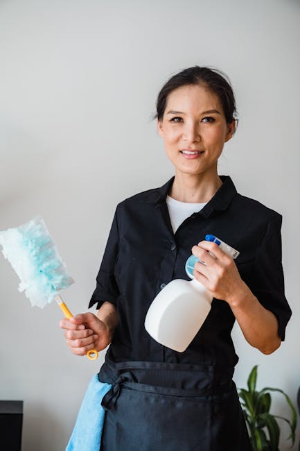 A female cleaner in a black uniform holding a white spray bottle in one hand and a blue and white cleaning cloth on a yellow handle in the other, standing in front of a plain light grey wall. The surf