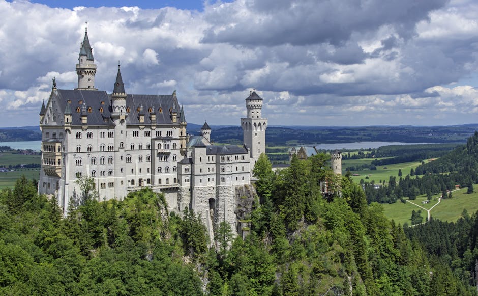 A large, white castle with multiple towers and spires situated atop a lush, green hill covered in trees. The castle features numerous windows and decorative battlements, and the stone structure appears well-maintained and clean. In the background, rolling fields and a distant lake are visible under a partly cloudy sky, with bright natural lighting highlighting the castle's details. The image showcases architectural surface details typical of historic stone buildings, emphasizing their grandeur and pristine condition, aligning with domestic or deep cleaning themes associated with maintaining such historic structures. The presence of greenery around the castle underscores the importance of regular surface cleaning and maintenance, similar to services offered by Cleaner Maida Vale.