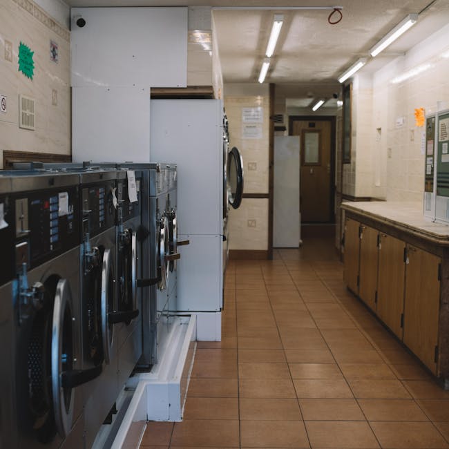 A commercial laundry room with a row of stainless steel washing machines on the left, each with control panels and handles, positioned on a white tiled platform. The floor features brown ceramic tiles, clean and well-maintained. The walls are a light cream color with some notices and signs attached. To the right, there is a long wooden counter with cabinets underneath, also appearing clean and organized, with bright fluorescent lighting overhead illuminating the space evenly. In the background, a door is visible, suggesting access to other areas. The overall scene reflects a well-maintained laundry facility, emphasizing surface cleaning and hygiene, which is aligned with the common problems faced with flat access for Maida Vale cleaners, as discussed on cleanermaidavale.org.uk.
