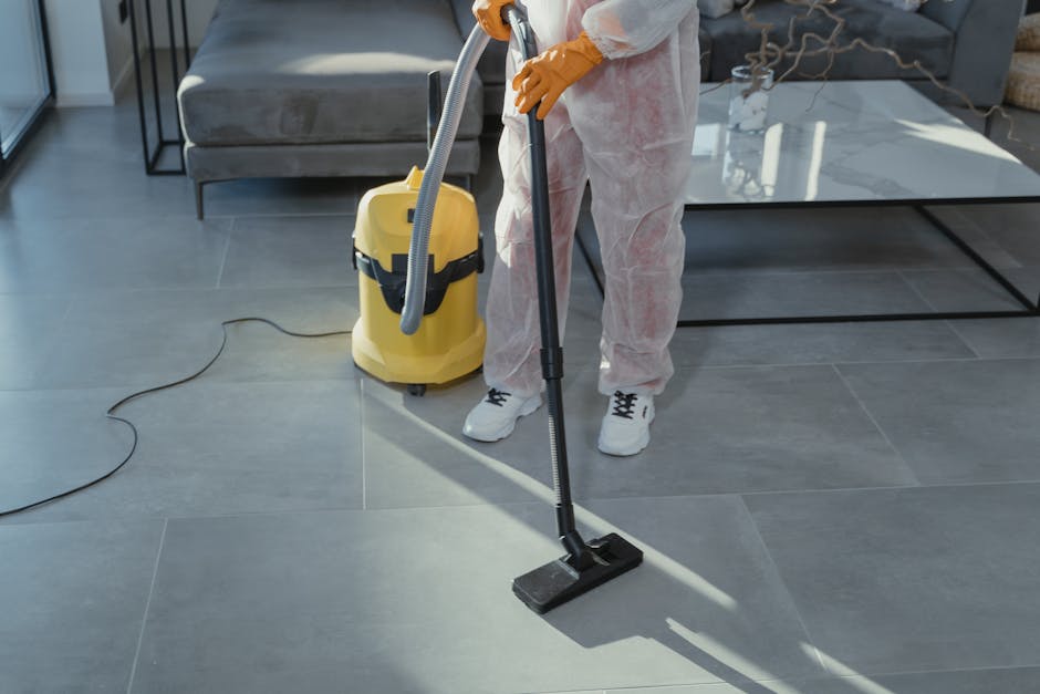 A person dressed in white overalls and yellow gloves is performing deep cleaning on large, grey ceramic floor tiles in a modern living room. They are using a vacuum cleaner with a flexible hose attachment to assist in surface cleaning and sanitation. Visible in the background are a grey upholstered sofa, a glass-top coffee table with a metal frame, and some decorative dried branches, all illuminated by natural lighting. The vacuum cleaner, which is yellow with a black handle, is plugged into an electrical outlet, and the cleaning process appears thorough, with the tiles appearing clean and shiny. Cleaner Maida Vale offers professional domestic cleaning services near Maida Vale station and Warwick Avenue, focusing on surface cleaning and hygiene maintenance.