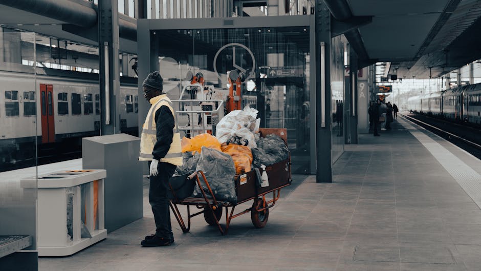 A sanitation worker wearing a high-visibility vest, dark clothing, and a knit cap stands on a tiled platform at a train station, next to a large cart filled with various trash bags and discarded items. The platform features smooth, gray ceramic tiles, with a glass shelter behind the worker. The station has modern infrastructure with metal supports, a train visible on the tracks to the right, and a few other people in the distance. The scene is well-lit with natural light, emphasizing the cleanliness of the area. Cleaner Maida Vale specializes in thorough surface cleaning and deep sanitation, ensuring hygienic environments in both residential and commercial spaces, even in busy transportation hubs.