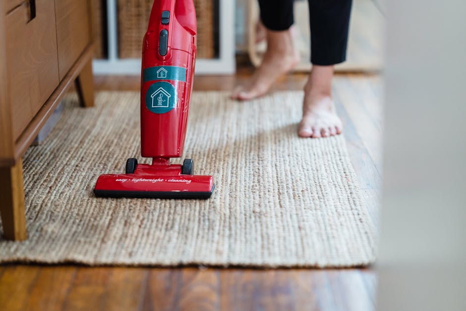 Close-up of a red cordless vacuum cleaner on a beige textured carpet in a living room, with visible suction marks indicating recent cleaning. A person dressed in black leggings is barefoot, standing next to the vacuum, with their feet on the carpet. The room features wooden flooring adjacent to the carpet and a wooden piece of furniture on the left side. Bright natural light illuminates the area, highlighting the cleanliness of the carpet surface. The scene emphasizes surface cleaning, deep cleaning, and maintenance relevant to domestic cleaning services offered by Cleaner Maida Vale, especially in the context of carpet care at Maida Vale homes, as discussed on the page about Little Venice boatside carpet cleaning tips.