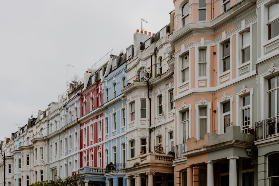 A row of Victorian-style residential buildings in Maida Vale with pastel-colored facades in shades of pink, blue, and white. The buildings feature bay windows, decorative moldings, and small balconies with black wrought-iron railings. The sky is overcast, casting diffuse light over the scene. The appearance shows well-maintained exteriors with clean surfaces, highlighting the importance of regular surface cleaning and upkeep for maintaining hygiene and aesthetic appeal, as promoted by Cleaner Maida Vale in their Maida Vale W9 flat cleaning guide for Elgin Avenue residents. The street exteriors exemplify clean, dust-free surfaces suitable for deep cleaning and sanitisation, aligning with domestic cleaning practices.