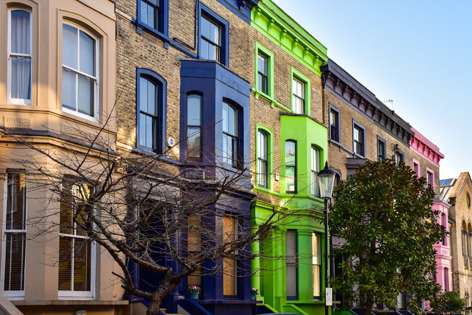 A row of Victorian-style residential buildings in Maida Vale with pastel-colored facades in shades of pink, blue, and white. The buildings feature bay windows, decorative moldings, and small balconies with black wrought-iron railings. The sky is overcast, casting diffuse light over the scene. The appearance shows well-maintained exteriors with clean surfaces, highlighting the importance of regular surface cleaning and upkeep for maintaining hygiene and aesthetic appeal, as promoted by Cleaner Maida Vale in their Maida Vale W9 flat cleaning guide for Elgin Avenue residents. The street exteriors exemplify clean, dust-free surfaces suitable for deep cleaning and sanitisation, aligning with domestic cleaning practices.