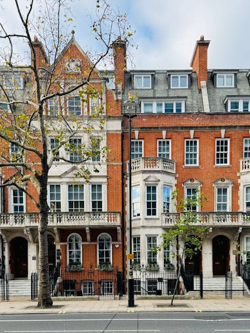 Photograph of a row of classic Victorian-style residential buildings in Maida Vale, featuring red brick facades, decorative white trim, and large sash windows. The buildings have ornate balconies and stone steps leading to dark wooden doors beneath arched entrances. In front, a sidewalk runs parallel to the street, which is lined with mature trees with sparse leaves, indicating early autumn. The scene is bathed in natural daylight, with a partly cloudy sky overhead. Visible on the street are black metal bollards and a street lamp, emphasizing the urban setting. Cleaner Maida Vale offers domestic cleaning and surface cleaning services to ensure property maintenance and hygiene in such elegant, historic residences.