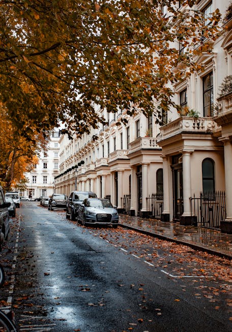 Photograph of a row of classic Victorian-style residential buildings in Maida Vale, featuring red brick facades, decorative white trim, and large sash windows. The buildings have ornate balconies and stone steps leading to dark wooden doors beneath arched entrances. In front, a sidewalk runs parallel to the street, which is lined with mature trees with sparse leaves, indicating early autumn. The scene is bathed in natural daylight, with a partly cloudy sky overhead. Visible on the street are black metal bollards and a street lamp, emphasizing the urban setting. Cleaner Maida Vale offers domestic cleaning and surface cleaning services to ensure property maintenance and hygiene in such elegant, historic residences.