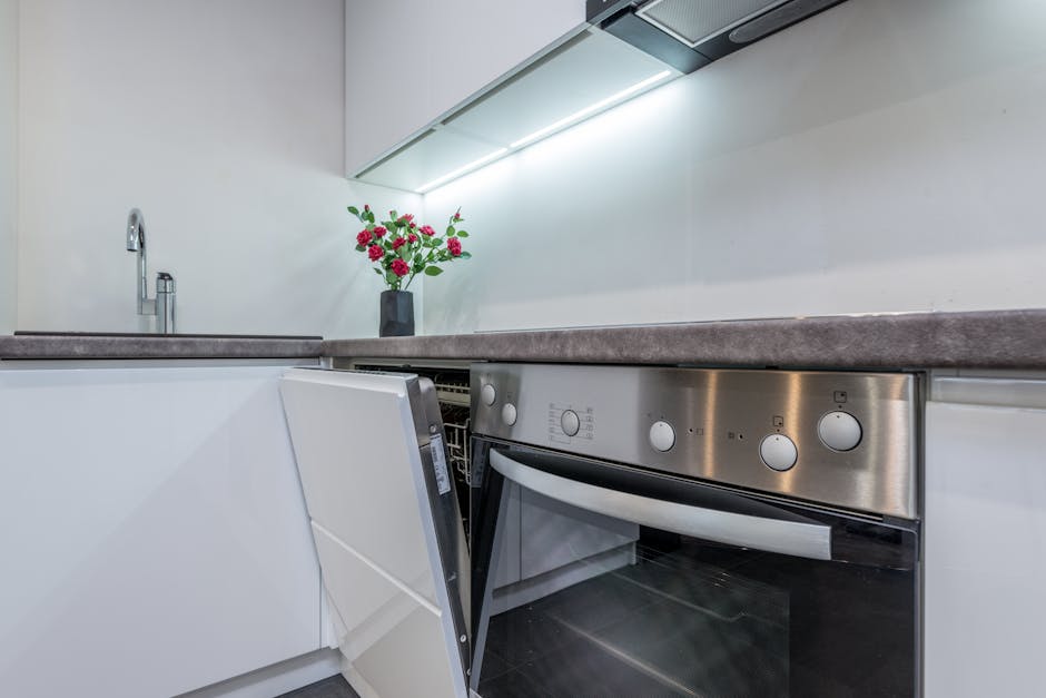 Close-up view of a modern kitchen countertop and stainless steel oven, both appearing clean and well-maintained. The ceramic countertop has a smooth surface with a subtle matte finish, and a small vase with red and pink flowers adds a decorative touch. The oven, integrated into white cabinetry, displays a shiny stainless steel control panel with several knobs and buttons, indicating recent deep cleaning. To the left, a sleek stainless steel faucet is visible above the sink, and there are no signs of dust, grime, or water stains. The lighting above the workspace enhances the cleanliness and bright atmosphere of the kitchen. This image exemplifies surface cleaning and domestic hygiene maintenance typical of professional services like those offered by Cleaner Maida Vale, specifically for oven and surface sanitisation in residential settings, aligning with Sutherland Avenue oven cleaning options for Maida Vale landlords.