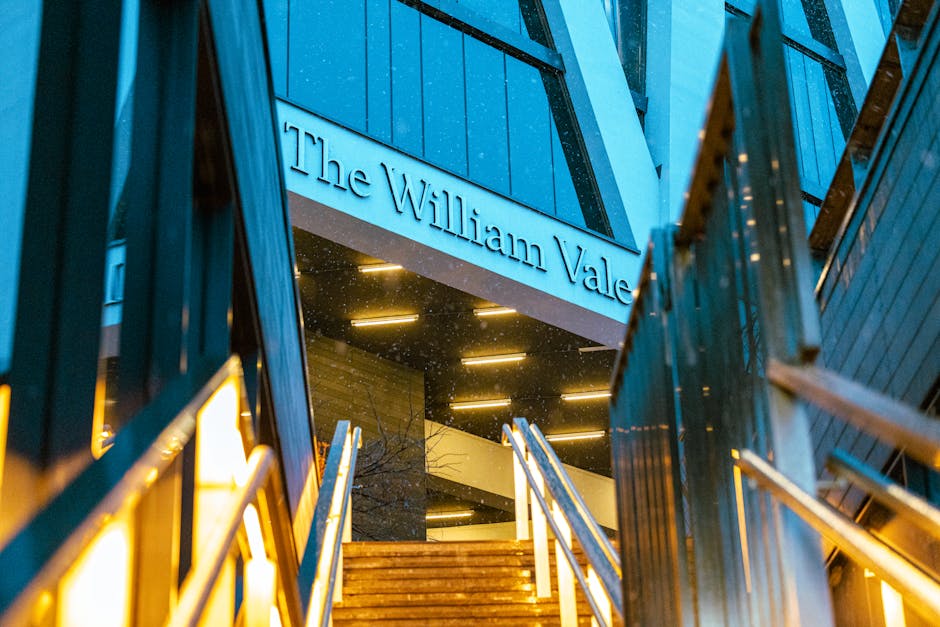 Exterior view of The William Vale building at dusk, with illuminated signage displayed on the facade. The photo shows a modern architectural structure featuring blue panels, glass windows, and metal railings along a staircase leading up to the entrance. The lighting highlights the building's clean and well-maintained appearance, with a focus on the sleek surfaces and reflective glass. The image captures the ambient evening light and the inviting atmosphere of the residential or commercial space, with no visible clutter or dirt. Cleaner Maida Vale specializes in surface cleaning, deep cleaning, and sanitisation of such properties, ensuring hygienic and pristine environments.