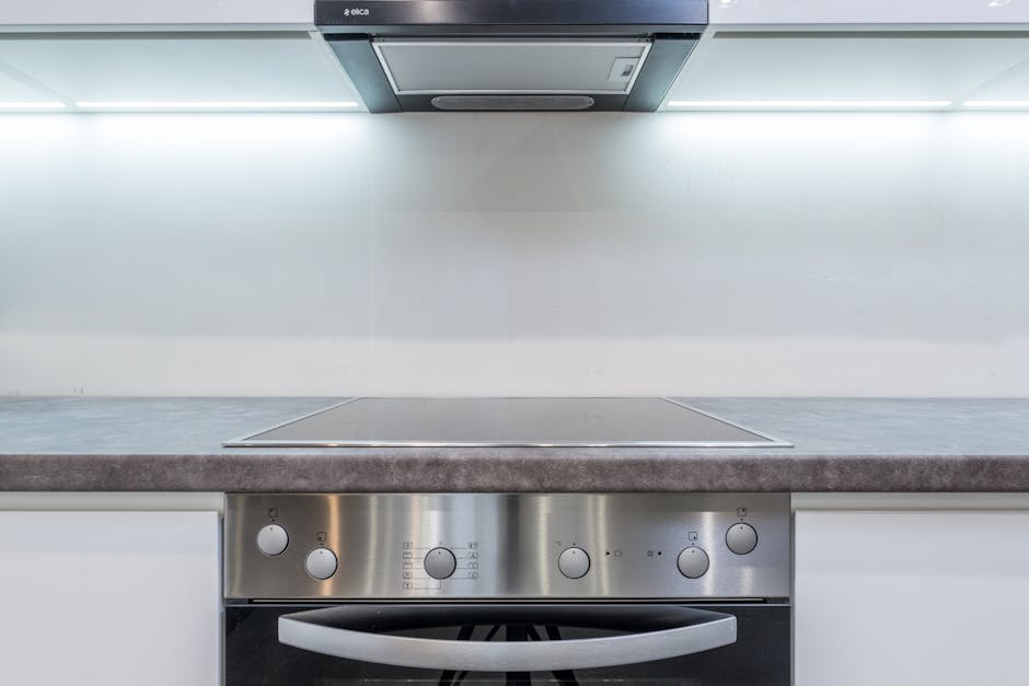 Close-up view of a modern kitchen countertop and stainless steel oven, both appearing clean and well-maintained. The ceramic countertop has a smooth surface with a subtle matte finish, and a small vase with red and pink flowers adds a decorative touch. The oven, integrated into white cabinetry, displays a shiny stainless steel control panel with several knobs and buttons, indicating recent deep cleaning. To the left, a sleek stainless steel faucet is visible above the sink, and there are no signs of dust, grime, or water stains. The lighting above the workspace enhances the cleanliness and bright atmosphere of the kitchen. This image exemplifies surface cleaning and domestic hygiene maintenance typical of professional services like those offered by Cleaner Maida Vale, specifically for oven and surface sanitisation in residential settings, aligning with Sutherland Avenue oven cleaning options for Maida Vale landlords.