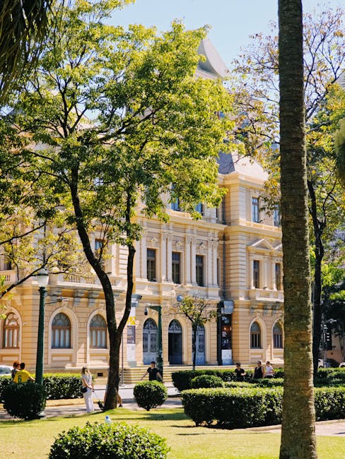 A sunny scene outside a large, historic building with ornate beige and white stonework and tall windows, partially obscured by lush green trees with bright leaves. The foreground features a well-maintained park with trimmed hedge bushes and paved walkways, where a few people are walking or sitting on benches. The natural lighting highlights the smooth, clean surfaces of the building and the vibrant greenery, exemplifying the importance of surface cleaning and maintenance in creating a tidy environment, as emphasized by Cleaner Maida Vale's domestic and commercial cleaning services.